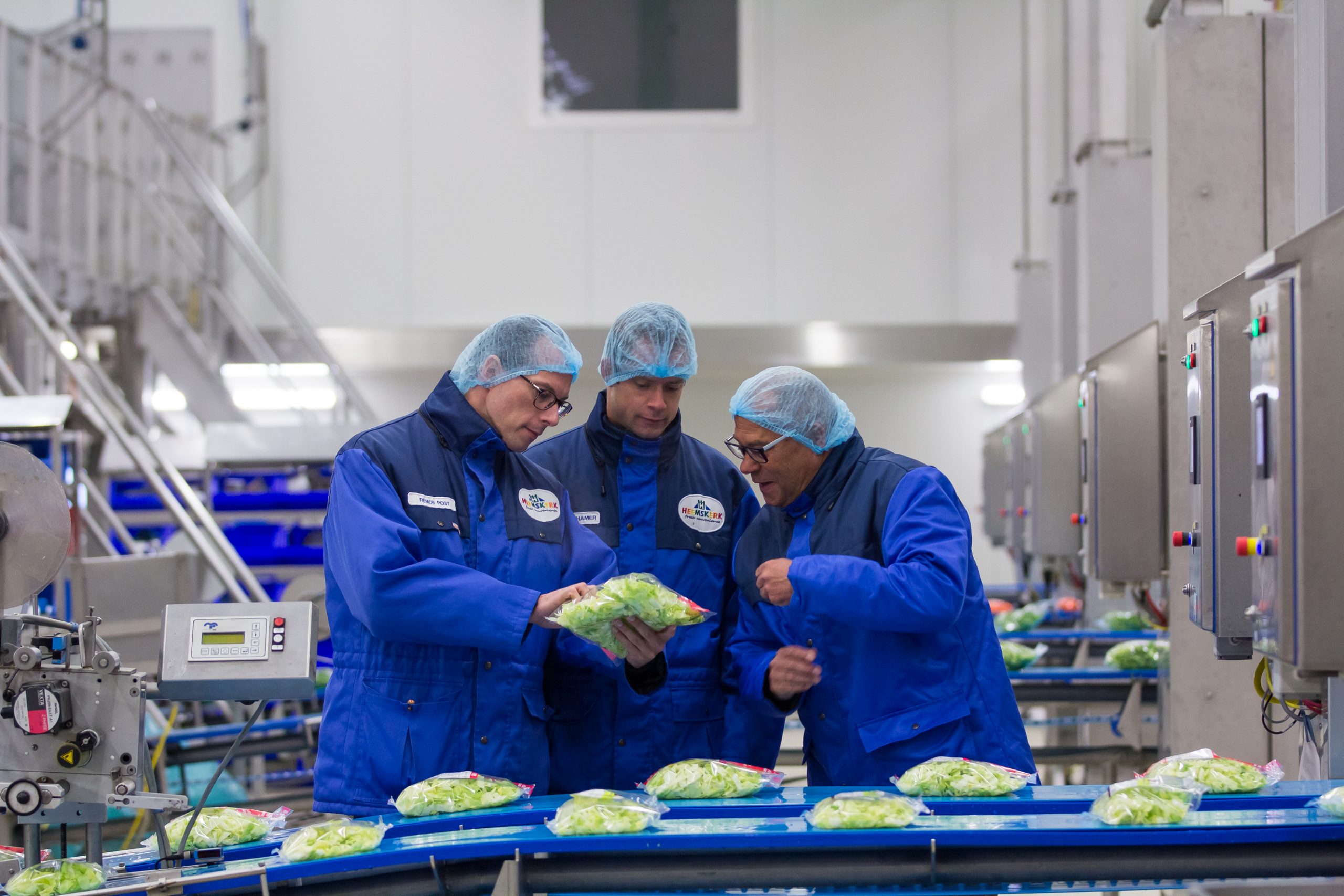 drie mannen staan in een productiehal in de food and beverage sector naast een lopende band met convenience sector producten en bekijken een verpakt product